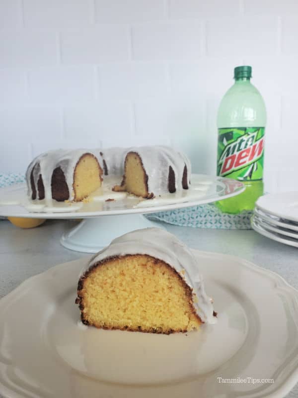 Slice of Mtn Dew cake on a white plate next to a platter with a Bundt Cake and a bottle of MTN Dew