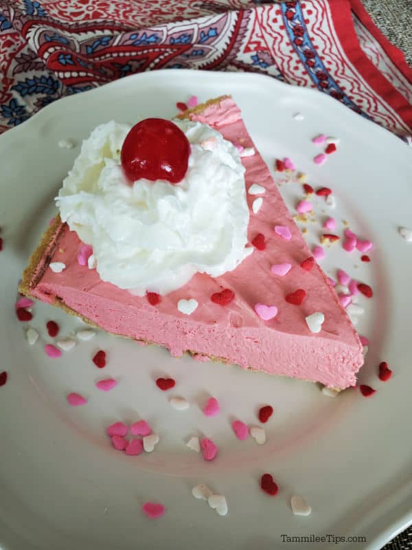 Pink Kool Aid Pie on a white plate garnished with heart sprinkles, whipped cream, and maraschino cherry