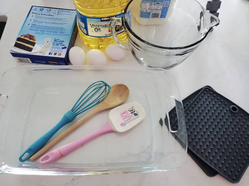 cake mix box, eggs, vegetable oil, glass batter bowl, whisk, wooden spoon, spatula, glass baking dish and pot holders on a white counter. 