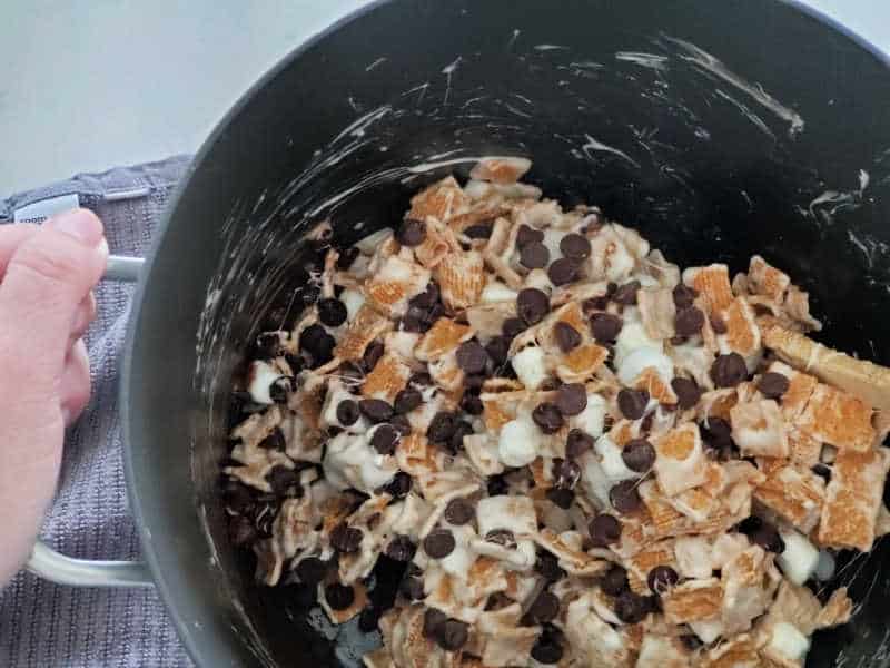 Golden grahams, marshmallows, chocolate chips stirred together with a wooden spoon in a pot