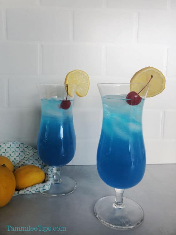 Blue lagoon drink in hurricane glasses with maraschino cherry and lemon wheel garnish. 