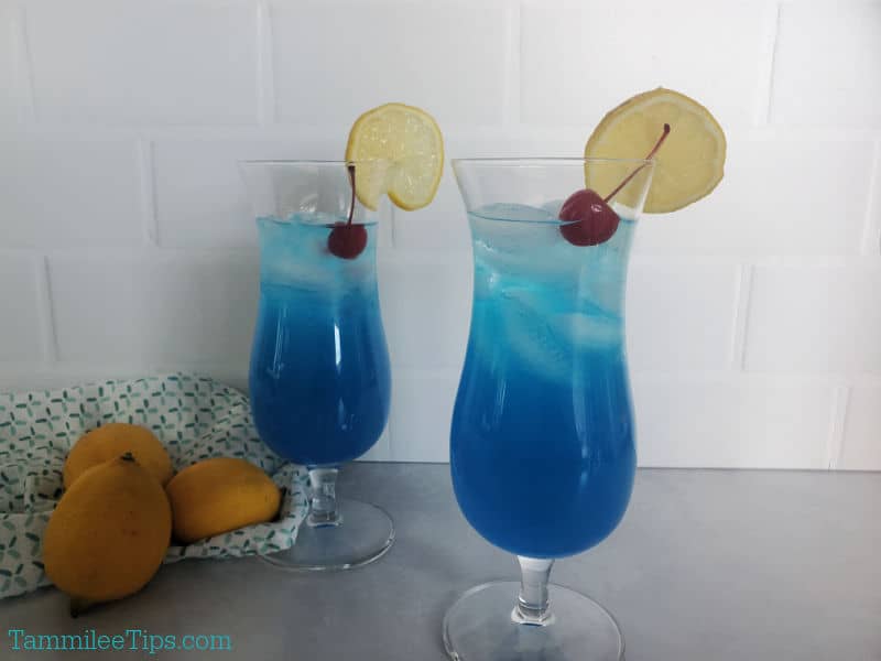 Two blue cocktails in hurricane glasses with maraschino cherry and lemon wheel garnish. 