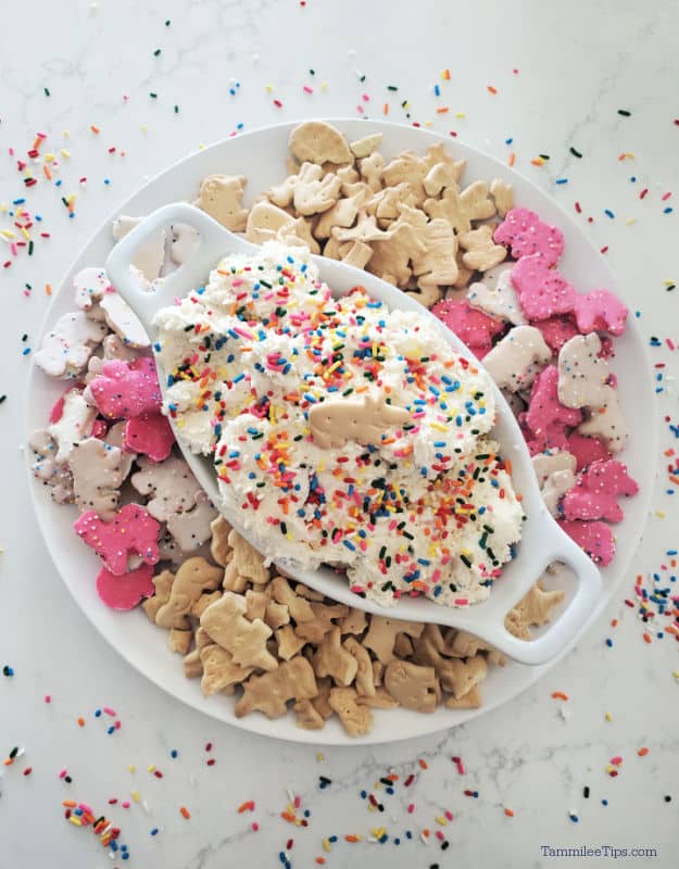 funfetti dip in a white bowl garnished with sprinkles and an animal cracker sitting on a white plate with additional animal crackers and cookies. 