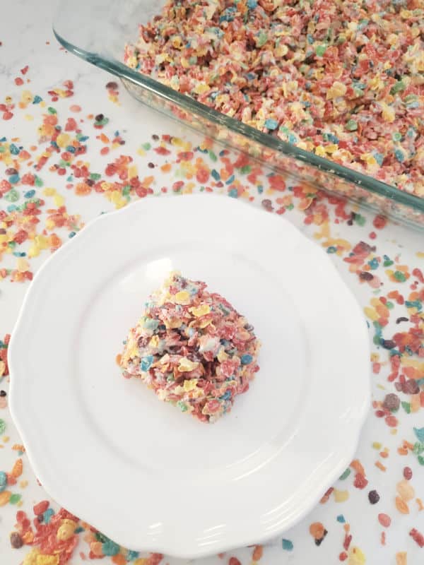 Fruity Pebbles Rice Crispy Treat on a white plate next to a glass baking dish with fruity pebbles treats. 