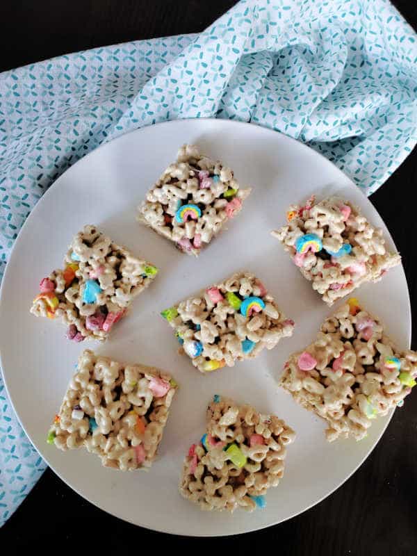 Squares of lucky charms treats spread around a white circle plate next to a cloth napkin 