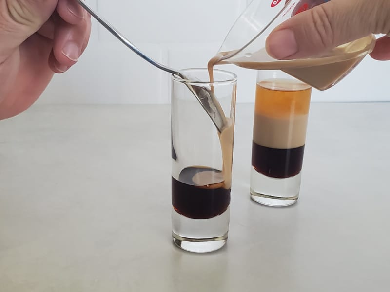 Irish Cream pouring over a layer of Kahlua in a shot glass next to a complete B52 Layered Shot