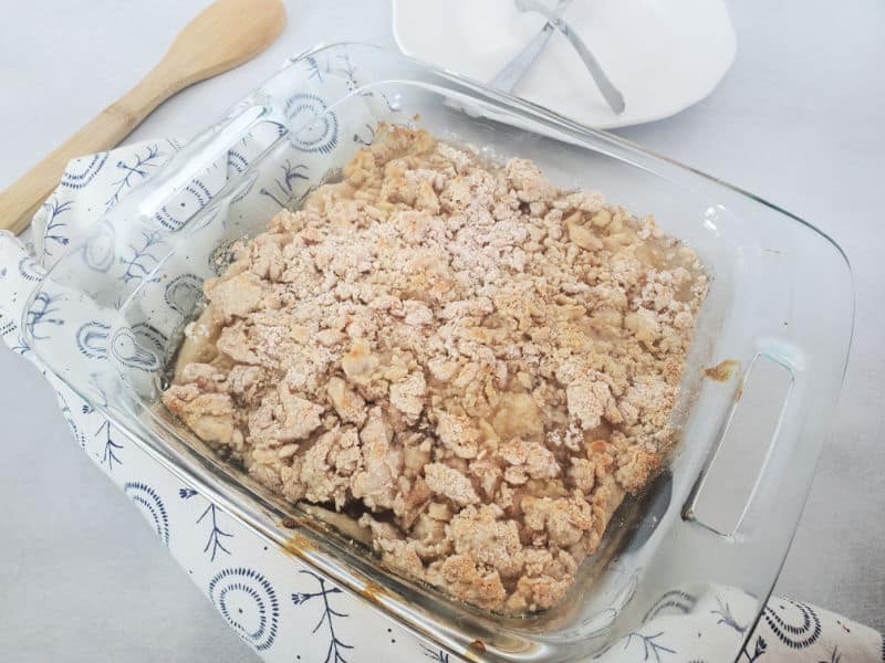 Baked Bisquick apple cobbler in a glass baking dish on a cloth napkin next to a wooden spoon and white plate with forks. 