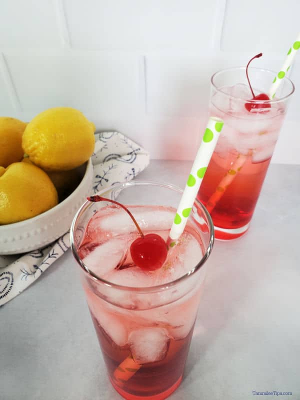 Looking down at two Dirty Shirley Drinks with maraschino cherry garnish and a paper straw next to a bowl of lemons. 