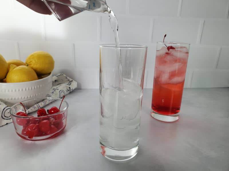 Bowl of lemons next to a glass of maraschino cherries next to a glass with clear liquid pouring into it and a red cocktail 