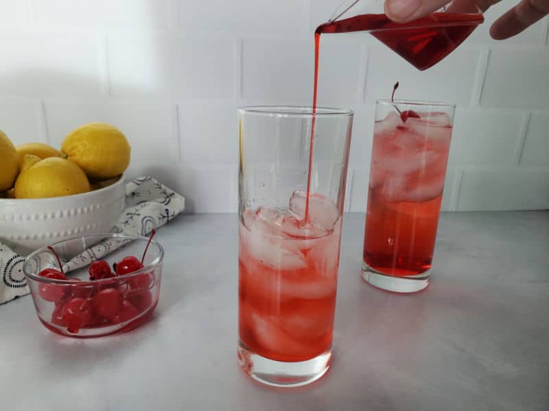 Red liquid pouring into a tall glass with ice next to a red drink, bowl of maraschino cherries, and a bowl of lemons. 