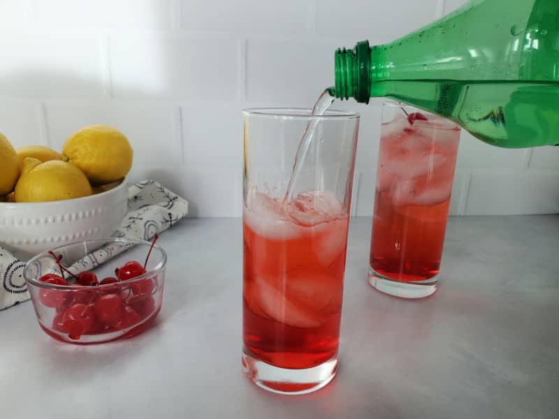 Sprite pouring into a red drink in a tall glass next to a bowl of maraschino cherries and a bowl of lemons. 