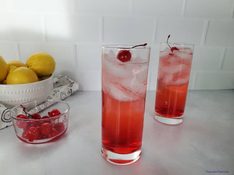 Two dirty Shirley drinks next to a bowl of maraschino cherries and a bowl of lemons