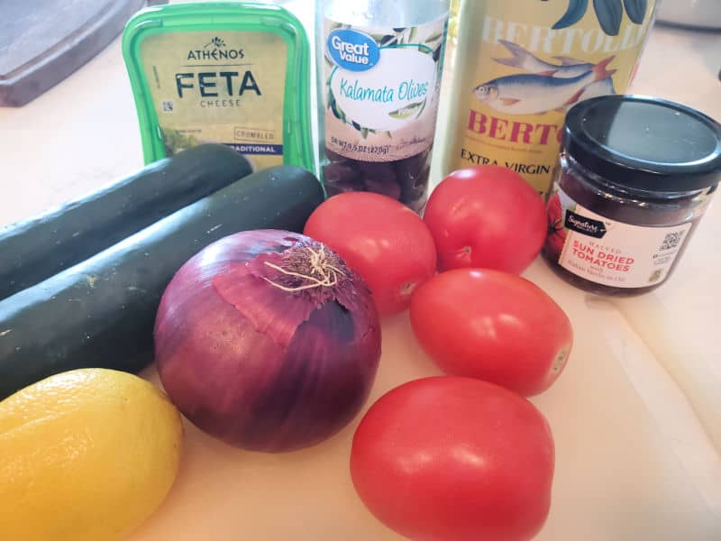 Feta, kalamata olives, olive oil, kalamata olives, cucumber, lemon, onions, and tomatoes on a cutting board. 