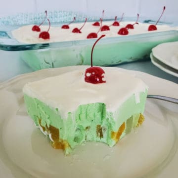 Square of green jello salad garnished with a cherry on a white plate next to a baking dish filled