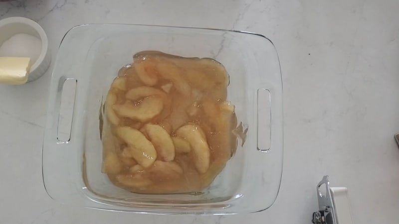 Apple pie filling in a glass baking dish next to a bowl with sugar and butter