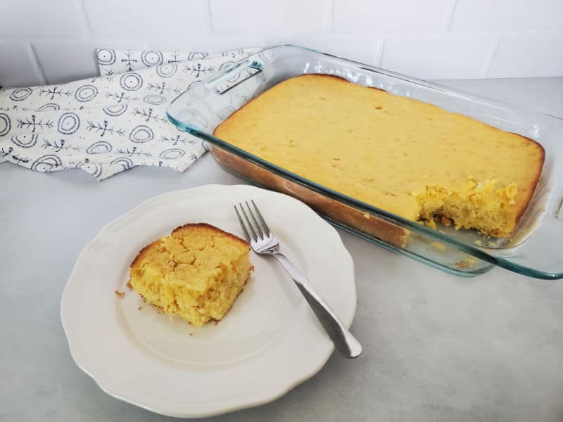 Square of cornbread on a white plate with a fork next to a glass baking dish with cornbread