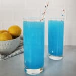 two bright blue kool aid slushies in tall glasses with paper straws, near a bowl of lemons
