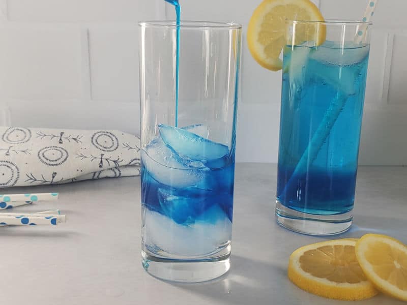Blue liquid pouring into a tall glass with ice next to a blue cocktail garnished with a lemon wheel