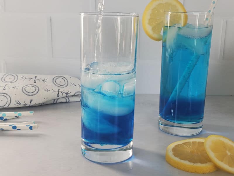 Clear liquid pouring into a glass with ice and blue liquid next to a glass with a blue cocktail and lemon wheel