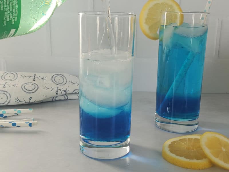 Clear liquid pouring from a Sprite bottle into a glass with blue liquid sitting next to a blue cocktail and lemon wheels