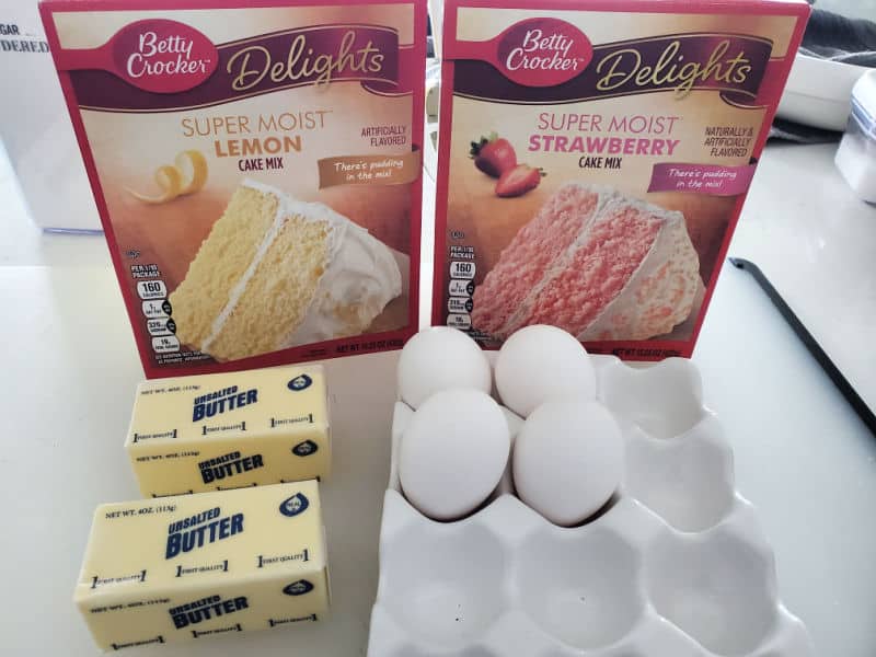 lemon cake mix, strawberry cake mix, butter, and eggs on a white counter. 