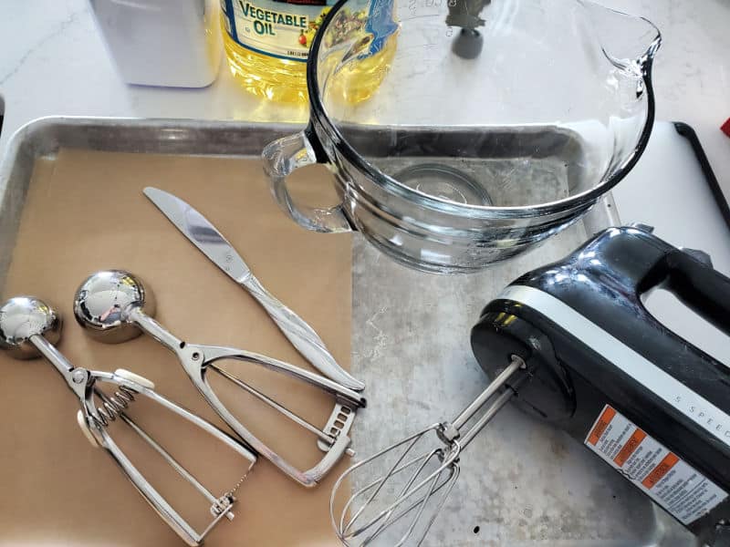 cookie scoops, knife, parchment paper, glass mixing bowl, hand mixer, and baking sheets on a counter. 