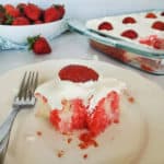square of strawberry poke cake with a strawberry garnish on a white plate with a fork, next to a glass cake pan and bowl of strawberries
