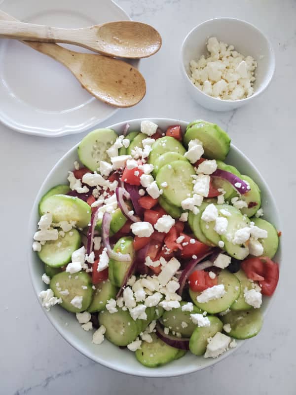 cucumber salad with feta in a white bowl next to a plate with two wooden spoons and a bowl of feta cheese