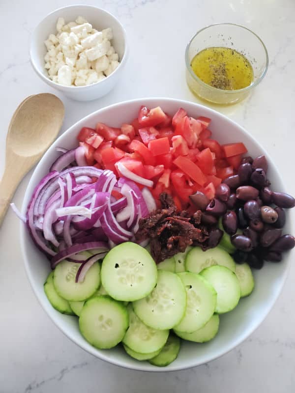 onions, tomatoes, kalamata olives, sundried tomatoes, and cucumbers in a white bowl next to a bowl of oil and a bowl of feta cheese