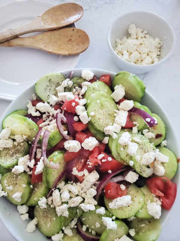 feta cheese topped cucumber salad next to wooden spoons and a bowl of feta cheese 