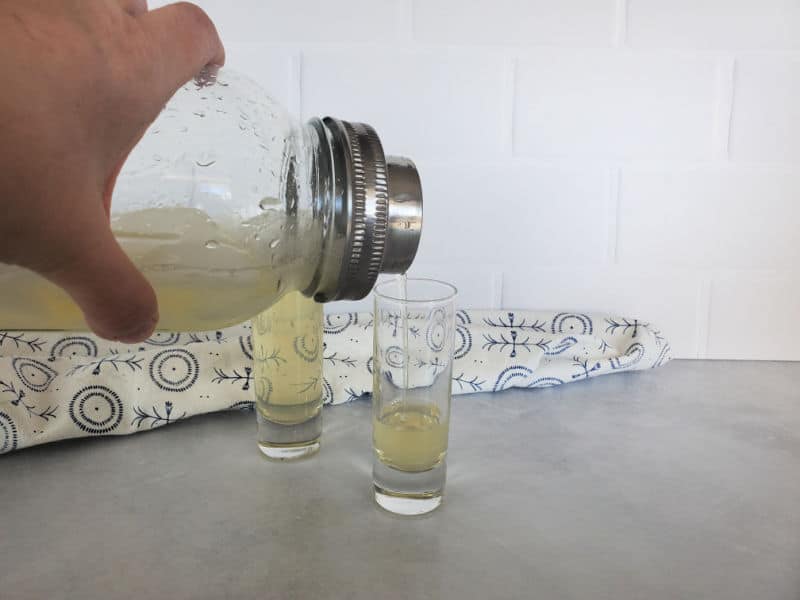 Clear glass mason jar cocktail shaker pouring a green tea shot into a shot glass with a cloth napkin behind 