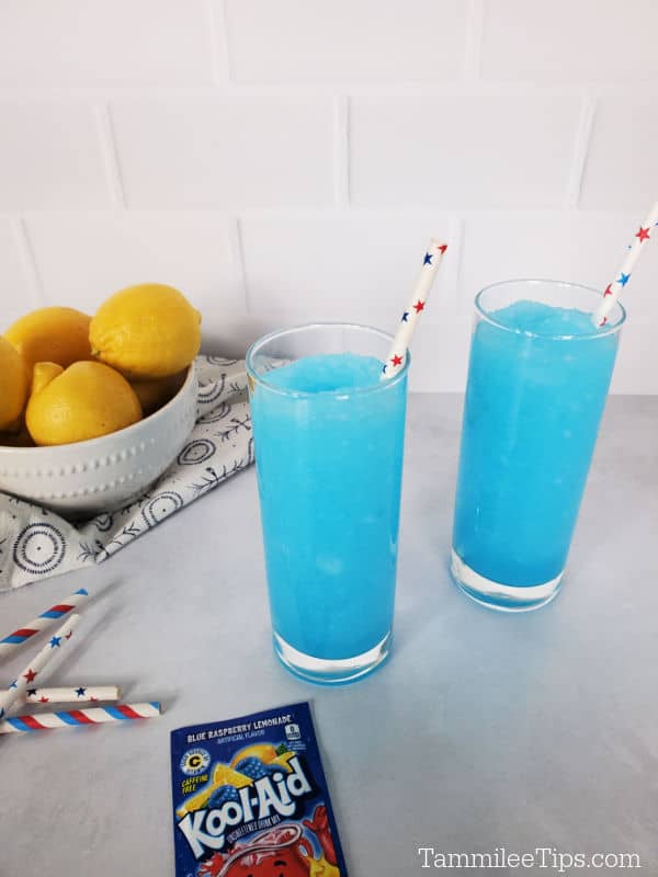 two bright blue kool aid slushies in tall glasses with paper straws, near a bowl of lemons and a package of Kool-Aid