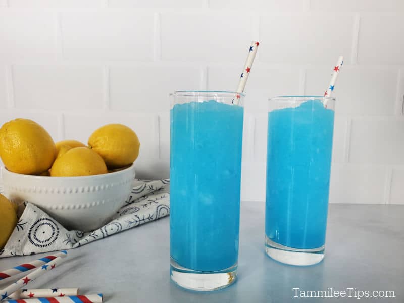 two bright blue kool aid slushies in tall glasses with paper straws, near a bowl of lemons and a stack of straws