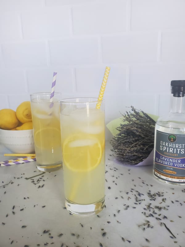 Lavender Lemonade Cocktails in tall glasses with lemon slices next to a bottle of Oakhurst Spirits Lavender Vodka and a bundle of Lavender. 