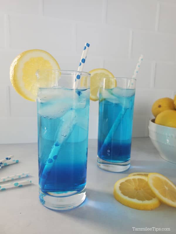 Two blue drinks in tall glasses garnished with lemon wheels and paper straws next to a bowl of lemons. 