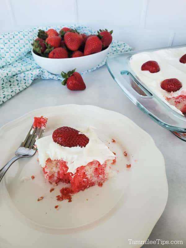 Slice of strawberry cake on a white plate with a fork next to a bowl of strawberries with a baking dish with cake in the corner