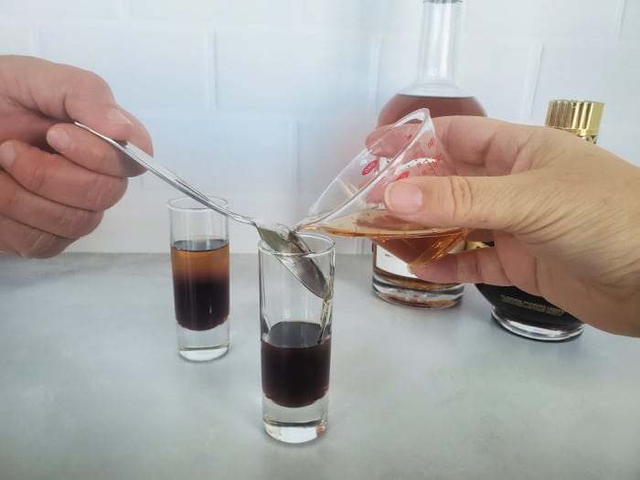 Hand holding a spoon over a partially filled shot glass while another hand pours over the back of the spoon