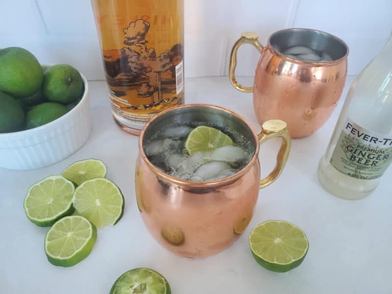 Kentucky Mule in a copper mug with lime slice, a bottle of bourbon, and a bottle of ginger beer. 