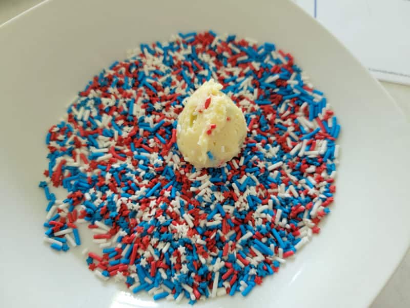 cookie dough ball sitting in red white and blue sprinkles on a white plate