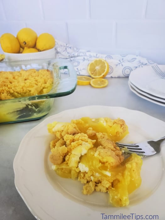 Serving of lemon dump cake on a white plate with the entire cake in the background with a bowl of lemons. 