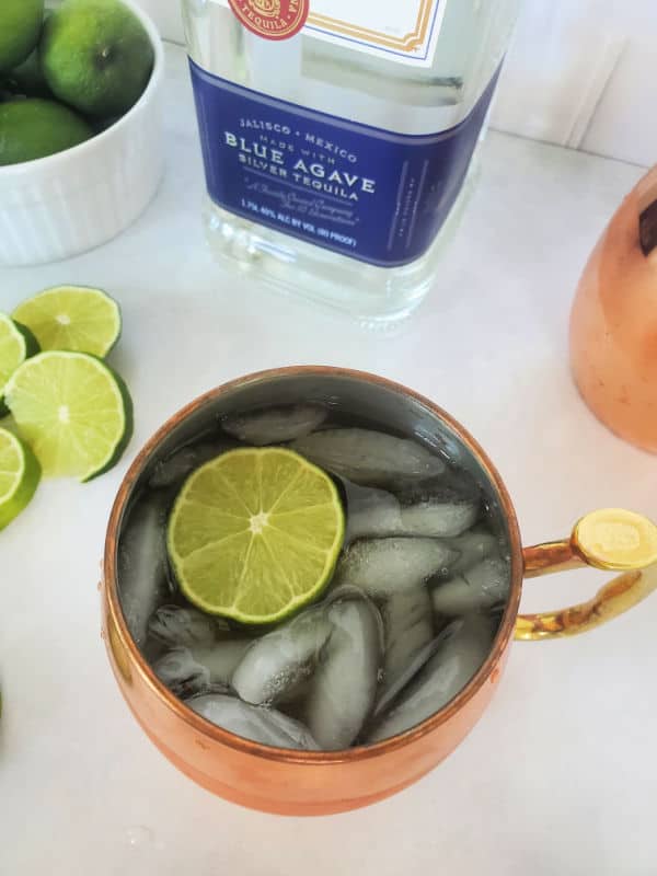 Mexican mule cocktail in a copper mug with a lime slice next to a bottle of tequila and additional lime slices