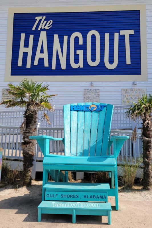 The Hangout printed on the side of the building above a blue Adirondack Chair with The Hangout Gulf Shores Alabama printed on it, next to palm trees