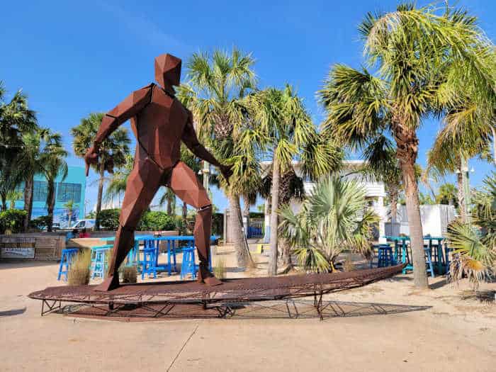 Large metal surfer statue on a metal surfboard next to palm trees