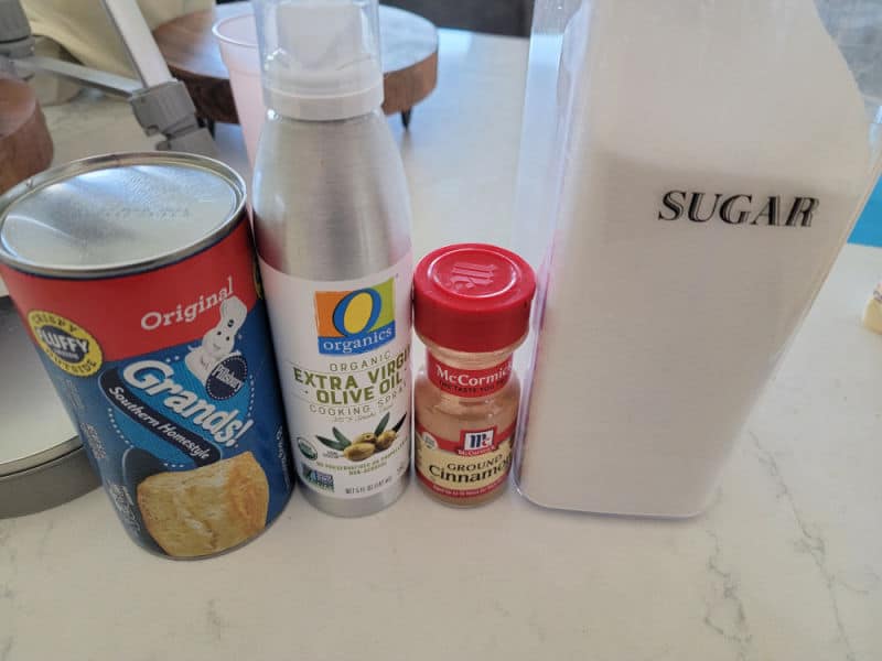 Air fryer Donuts ingredients, Grands Biscuits, Olive Oil Spray, Cinnamon, and Sugar on a white counter. 