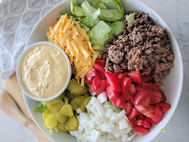 Big Mac Salad in a white bowl with big mac sauce, dill pickles, onions, tomatoes, ground beef, lettuce, and shredded cheddar cheese in a white bowl with wooden spoons and a cloth napkin