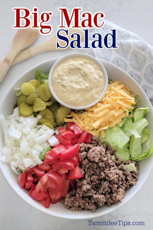 Big Mac Salad text over a Big Mac Salad in a white bowl with big mac sauce, dill pickles, onions, tomatoes, ground beef, lettuce, and shredded cheddar cheese in a white bowl with wooden spoons. 