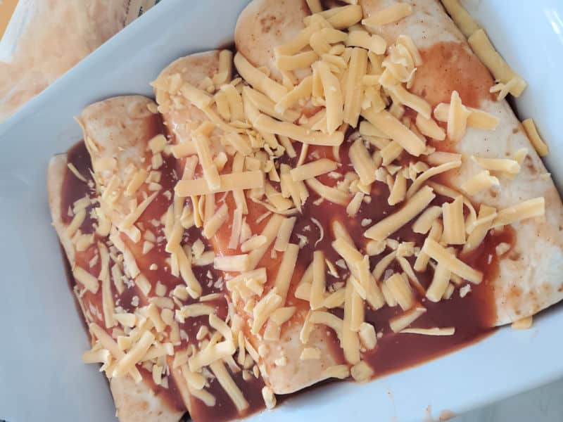 Shredded cheese covering tortillas and enchilada sauce in a white baking dish. 