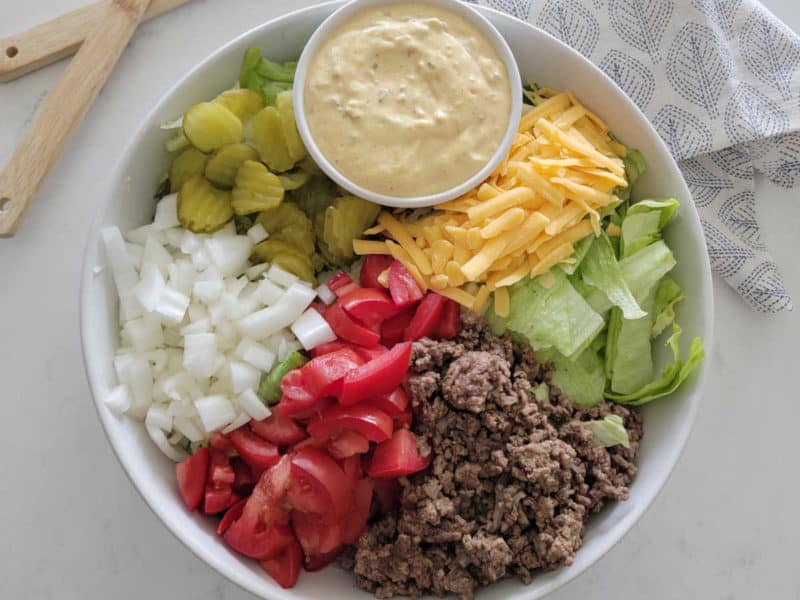 Big Mac Salad in a white bowl with big mac sauce, dill pickles, onions, tomatoes, ground beef, lettuce, and shredded cheddar cheese. 