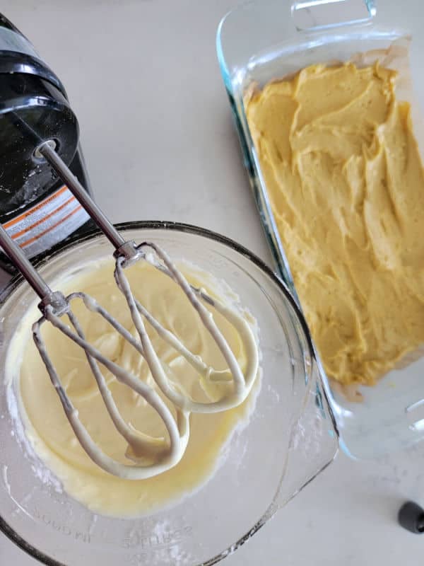 Handheld mixer above a bowl of cream cheese topping next to the base layer of ooey gooey butter cake