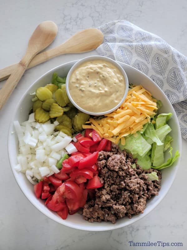 Big Mac Salad in a white bowl with big mac sauce, dill pickles, onions, tomatoes, ground beef, lettuce, and shredded cheddar cheese. in a white bowl with wooden spoons